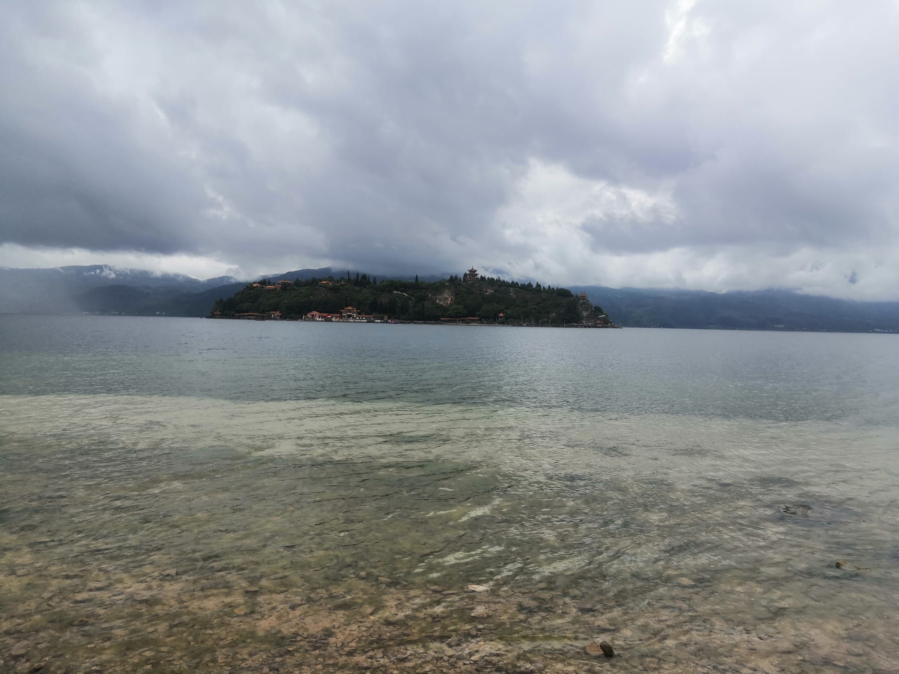 Clear shallows over a pebbled lakebed at Fuxian Lake, with Gushan island beyond under heavy cloud