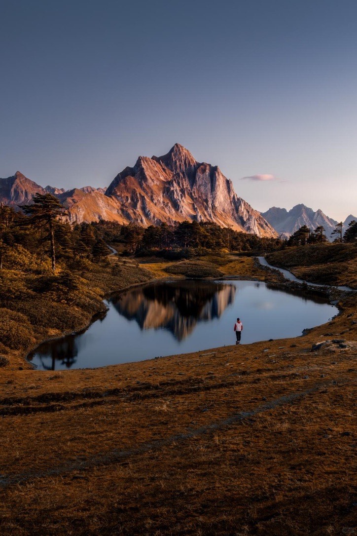 Lone hiker beside an alpine tarn reflecting a golden sunset-lit peak near Shangri-La