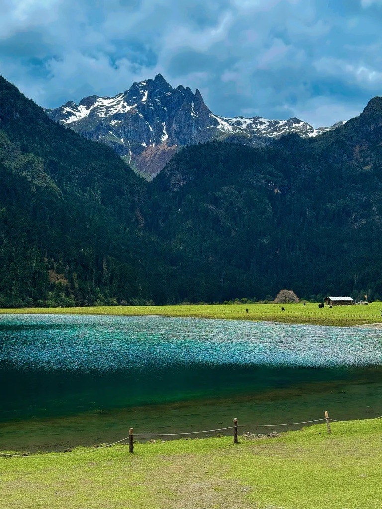 Mountain landscape with snow-capped peaks and a turquoise Wudi lake in Shangri-La. Green grass and a small cabin in the foreground under a cloudy sky.