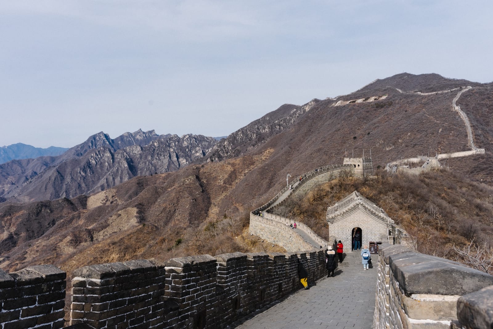 Restored ramparts at Mutianyu in winter — visitors on the wide paved walkway, the wall climbing the bare ridge to watchtowers above