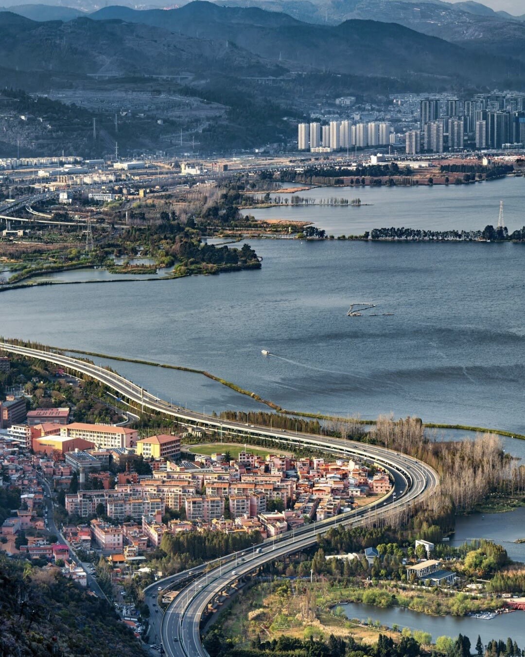 Aerial view of a broad Central Yunnan lake edged by a curving elevated highway, town and distant mountains