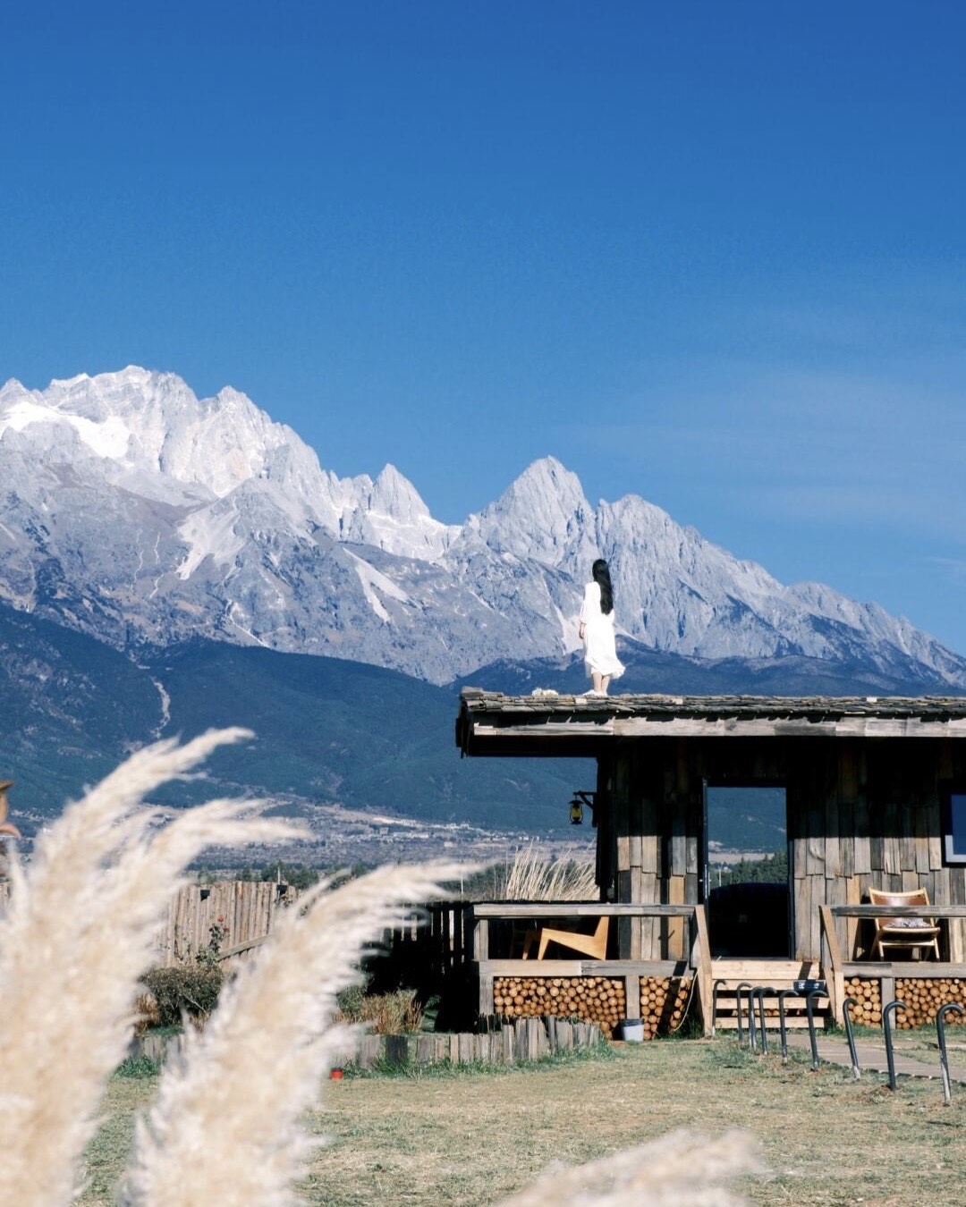 Woman in white on a wooden lodge roof facing snow-capped Lijiang peaks, northwest Yunnan