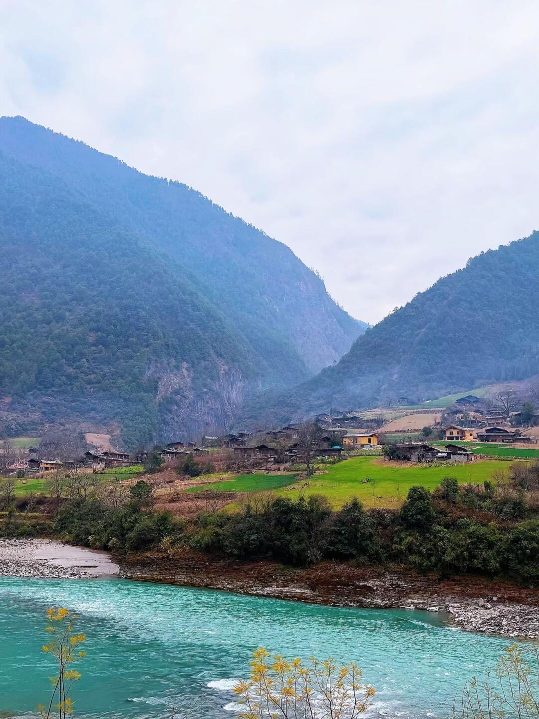 Turquoise Nujiang river winding past a riverside village and green fields below forested gorge peaks