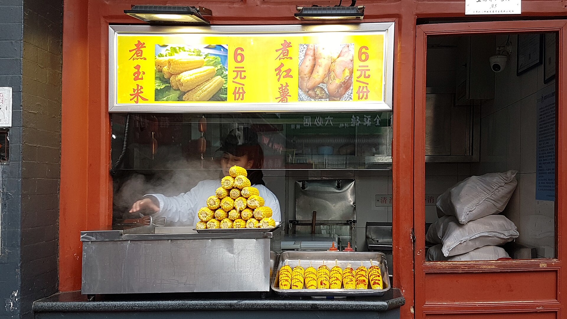 A vendor stacking a glistening golden pyramid of steamed corn (玉米) amid rising steam at a Beijing street-food stall