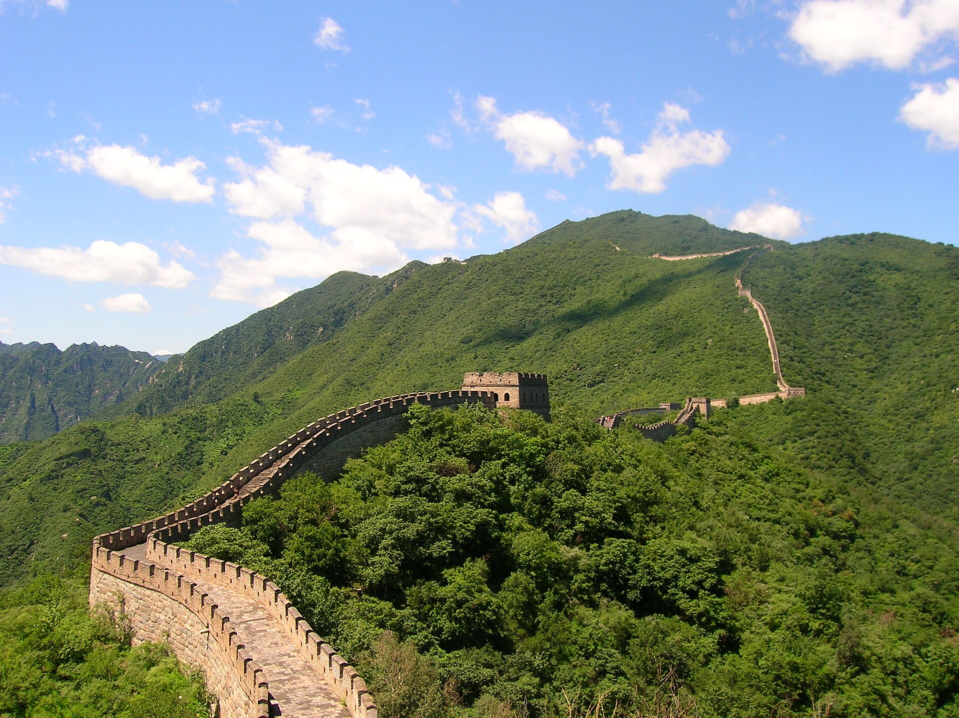 The Great Wall snaking along a densely forested ridgeline under a blue summer sky near Beijing, a watchtower at the saddle