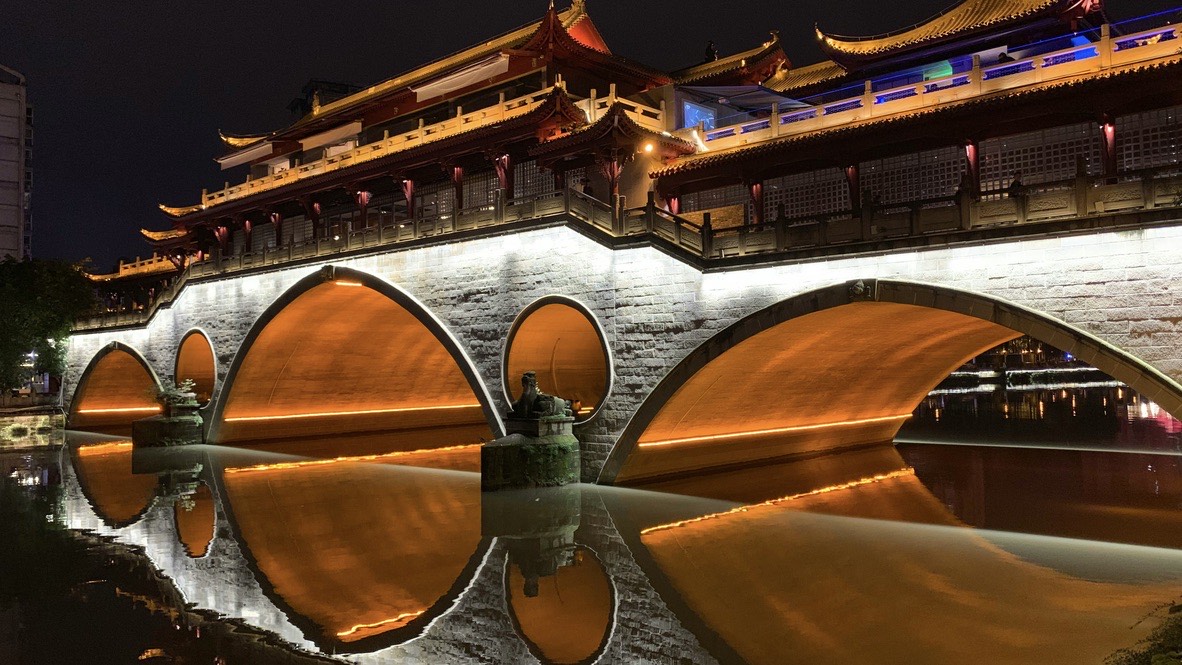 Illuminated tiered wooden pavilion atop a stone arch bridge in Chengdu, its lit arches mirrored in the calm river at night.