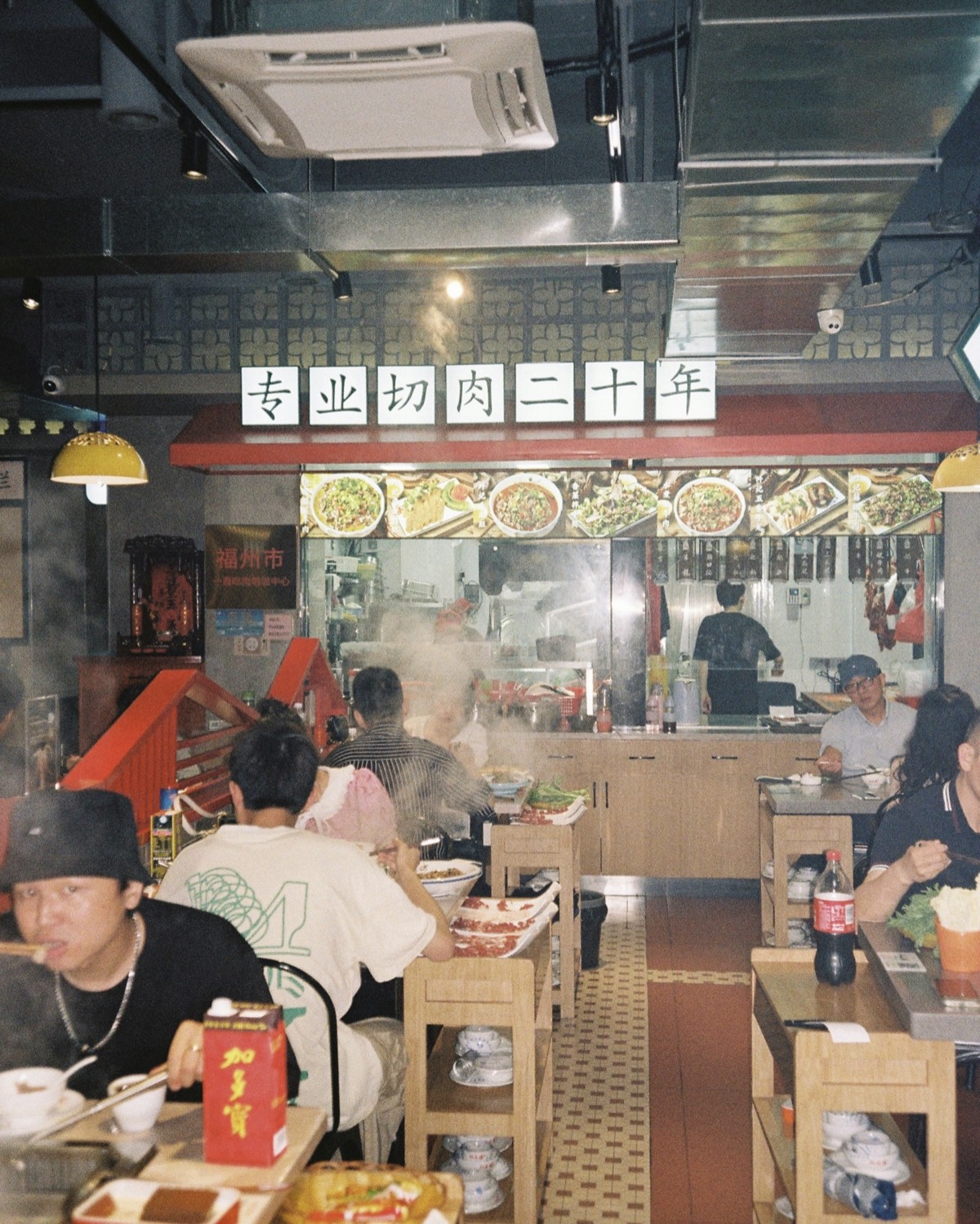 Busy Chengdu restaurant with diners at wooden tables, a steamy open kitchen and photo menu on the back wall.