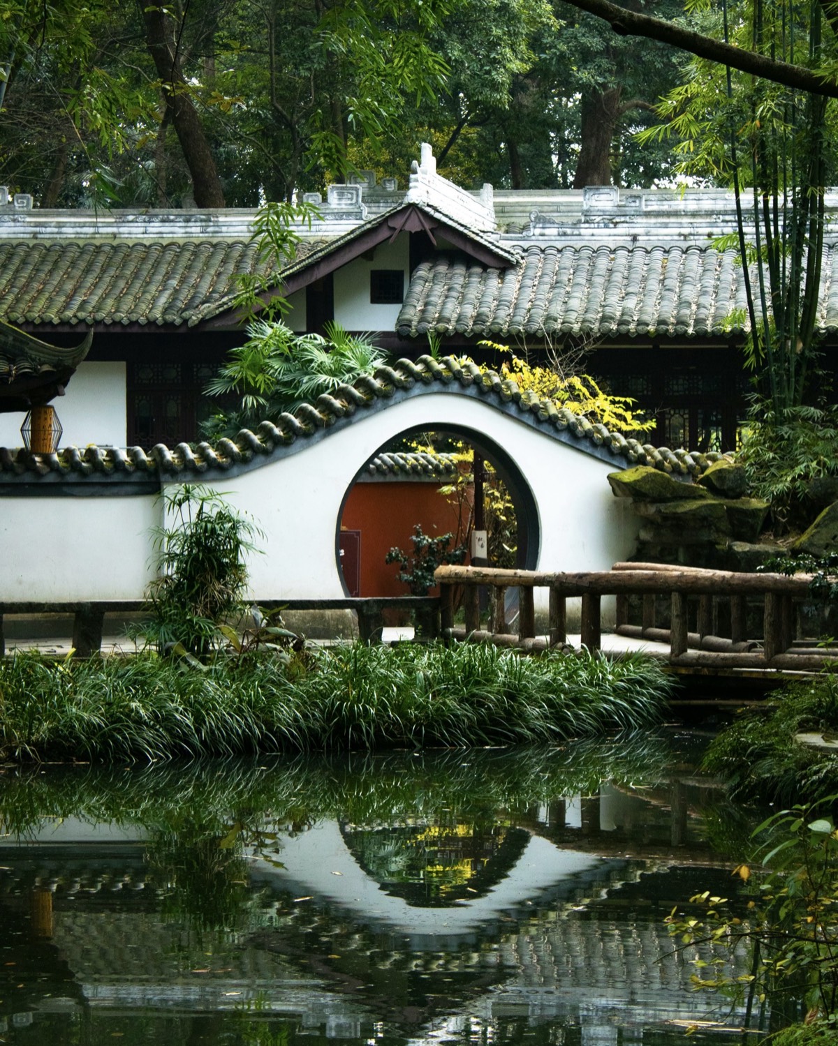 White-walled pavilion with a circular moon gate and grey tiled roofs reflected in a still garden pond in Chengdu.