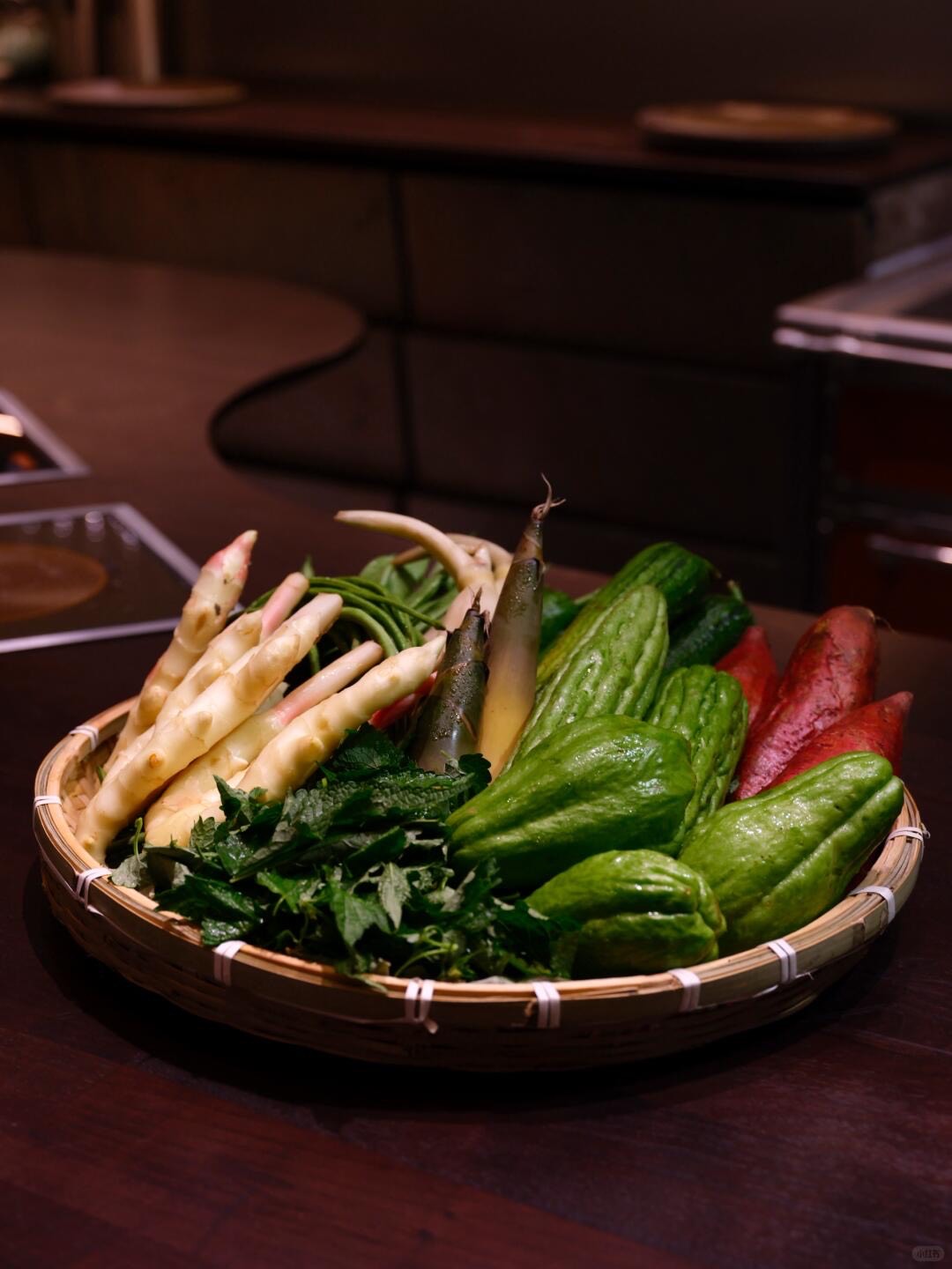 Bamboo basket of fresh Sichuan vegetables including bamboo shoots, chayote and greens on a dark restaurant table.