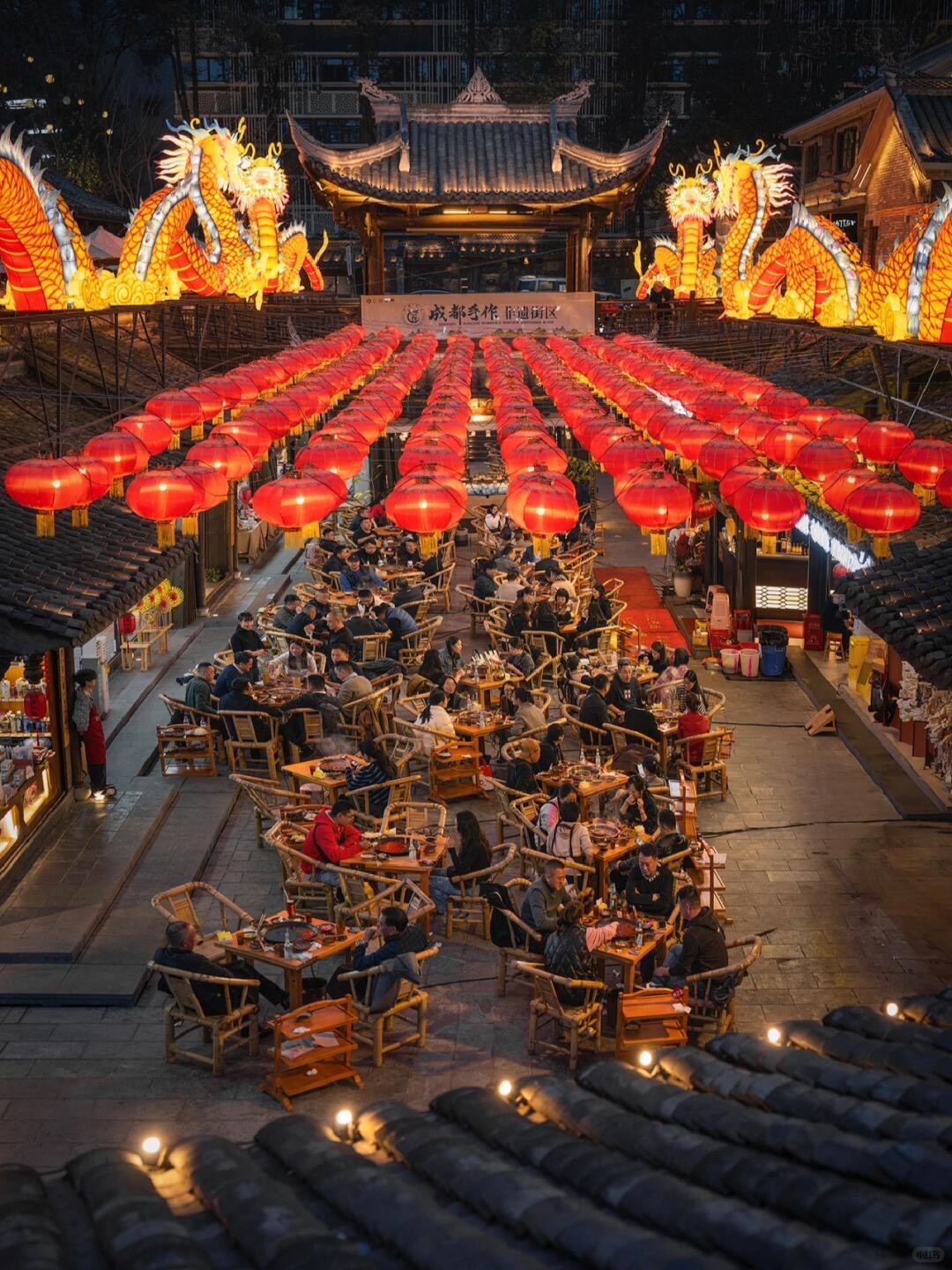 Chengdu night market lit by rows of red lanterns and glowing dragon displays, with diners at bamboo tables below.