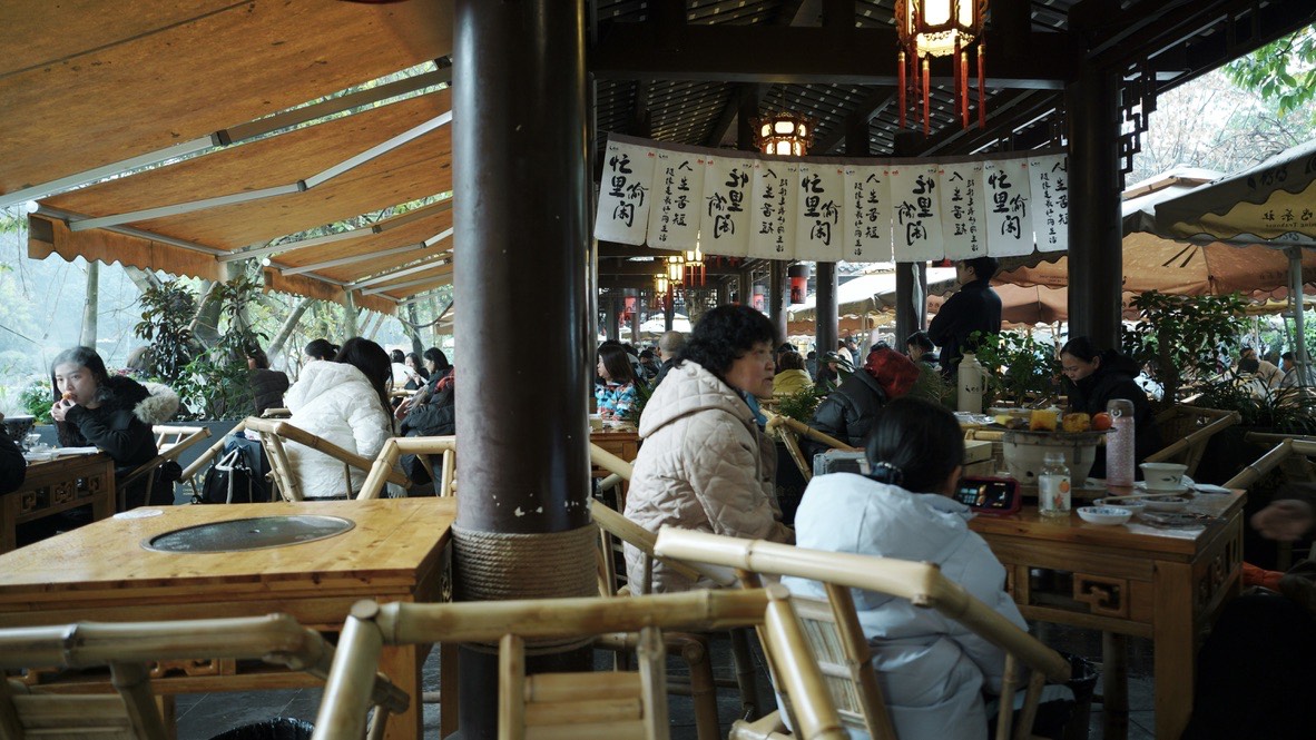 Guests relax in bamboo chairs under a covered pavilion at a bustling open-air Chengdu teahouse hung with calligraphy banners.