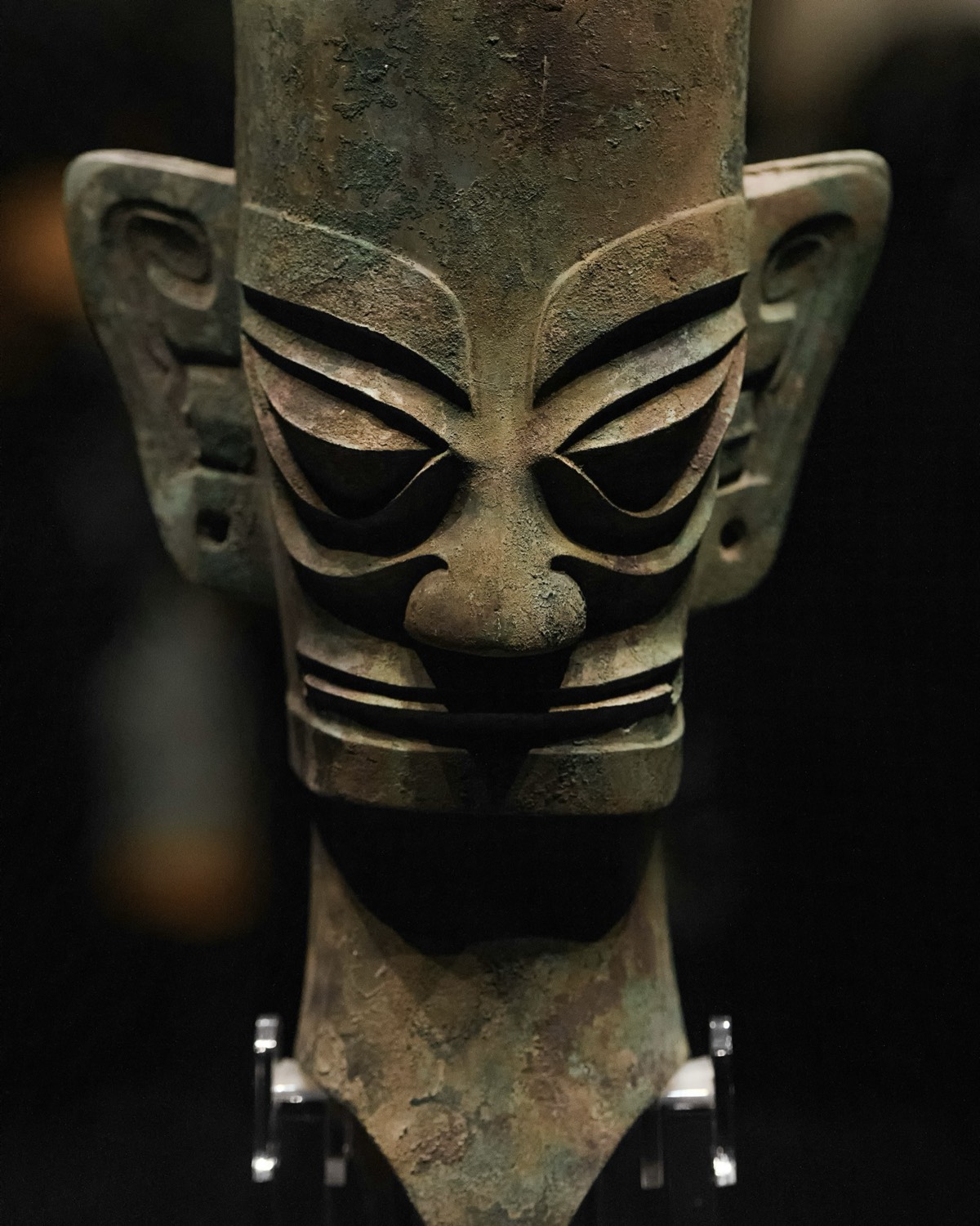 Ancient bronze Sanxingdui head with angular eyes and large ears displayed against a dark museum backdrop near Chengdu.