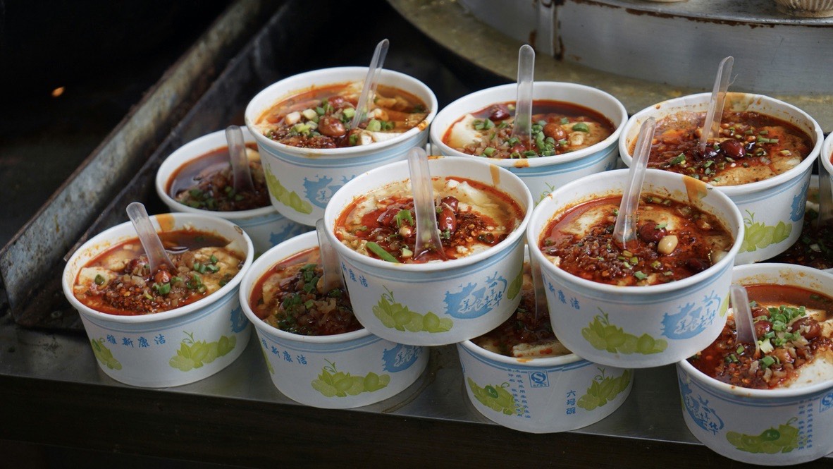 Rows of takeaway bowls of spicy tofu topped with chilli oil, peanuts and spring onion at a Chengdu street stall.