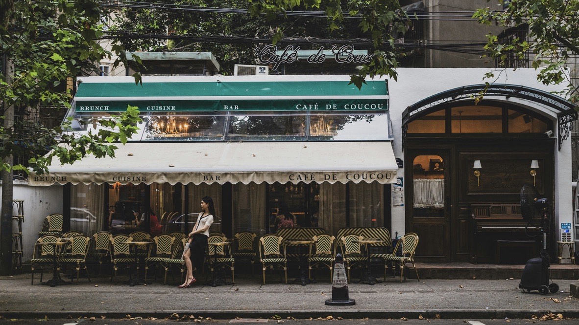 Leafy street facade of a Chengdu cafe with a green awning and rows of woven rattan bistro chairs along the pavement.