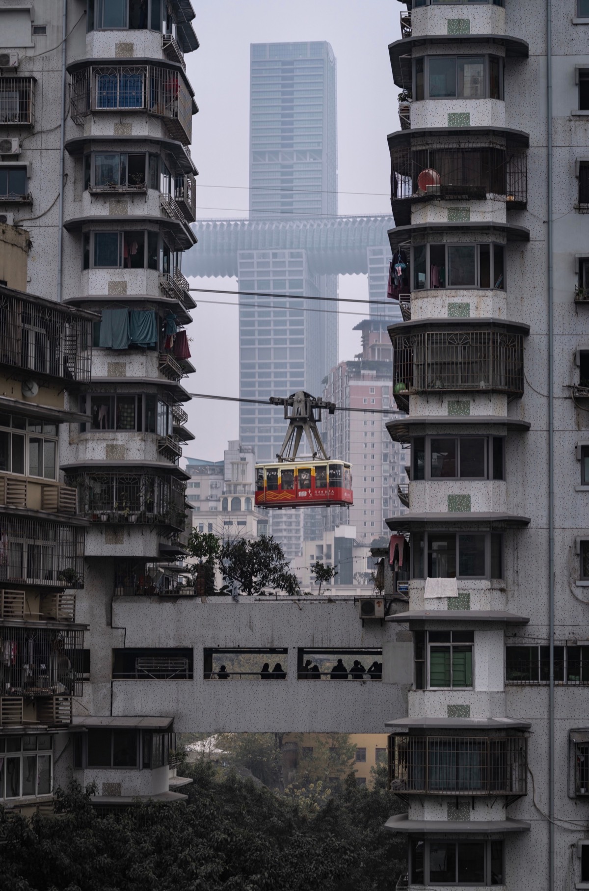 Red and yellow river cableway cabin crossing between old apartment towers with a hazy Chongqing skyscraper behind.