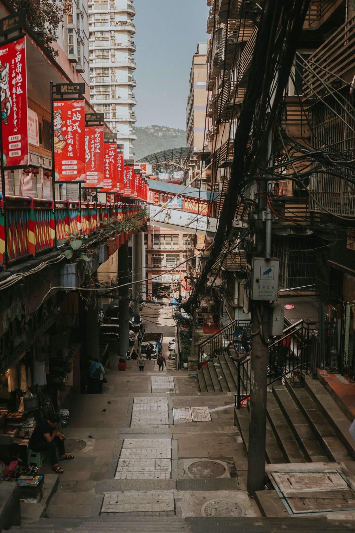 Steep tiled staircase alley in Chongqing lined with red shop banners, tangled wires and high-rise towers behind.