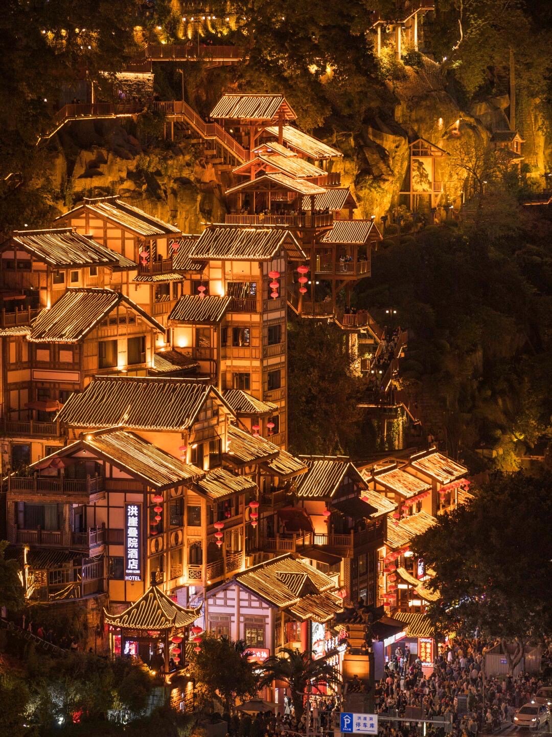 Golden-lit tiered stilt houses and hanging lanterns rising up a cliffside in Chongqing's old town at night.