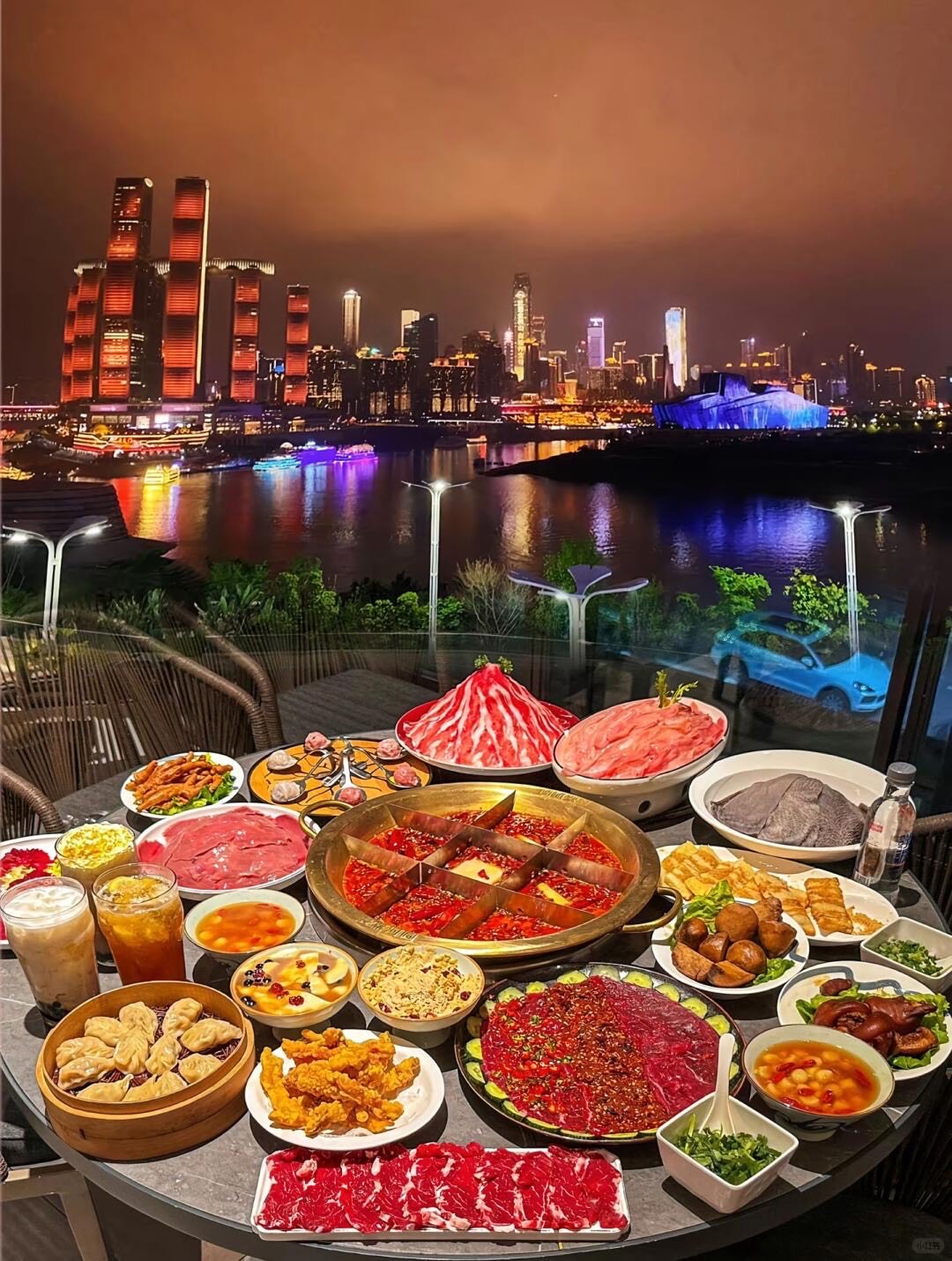 Lavish spread of Chongqing hotpot and sliced meats on a terrace overlooking the glowing riverside night skyline.