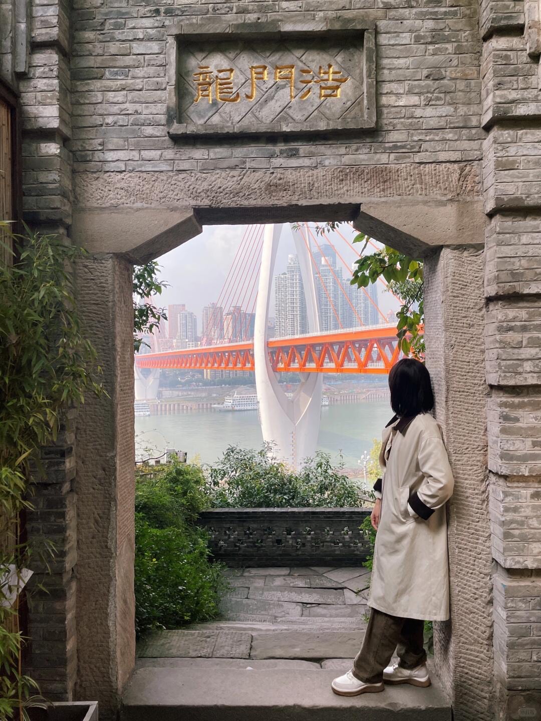 Woman by a stone brick gateway framing a view of the red arch bridge and river skyline in Chongqing's Longmenhao.