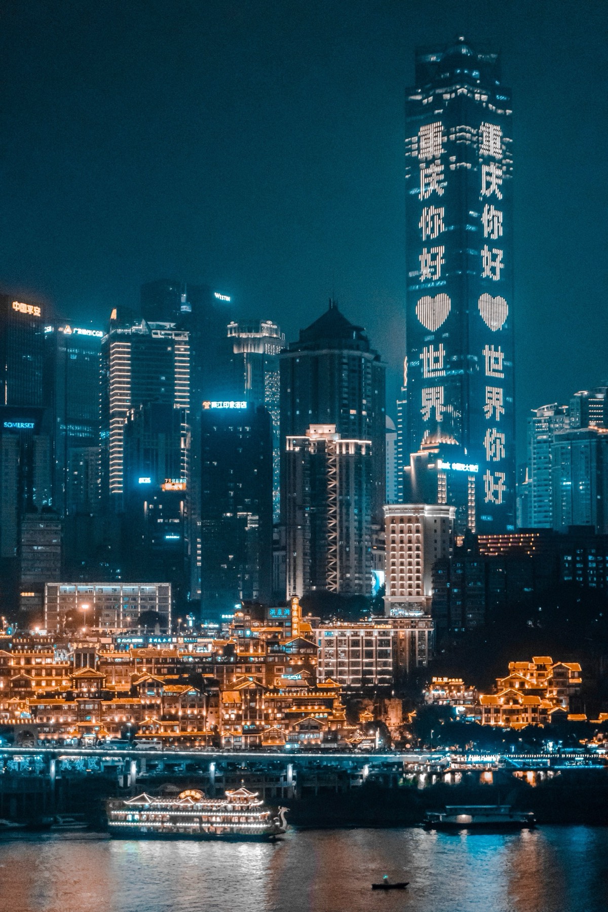Chongqing riverfront skyline at night with a skyscraper lit in glowing characters above golden waterfront buildings and a river cruise boat.