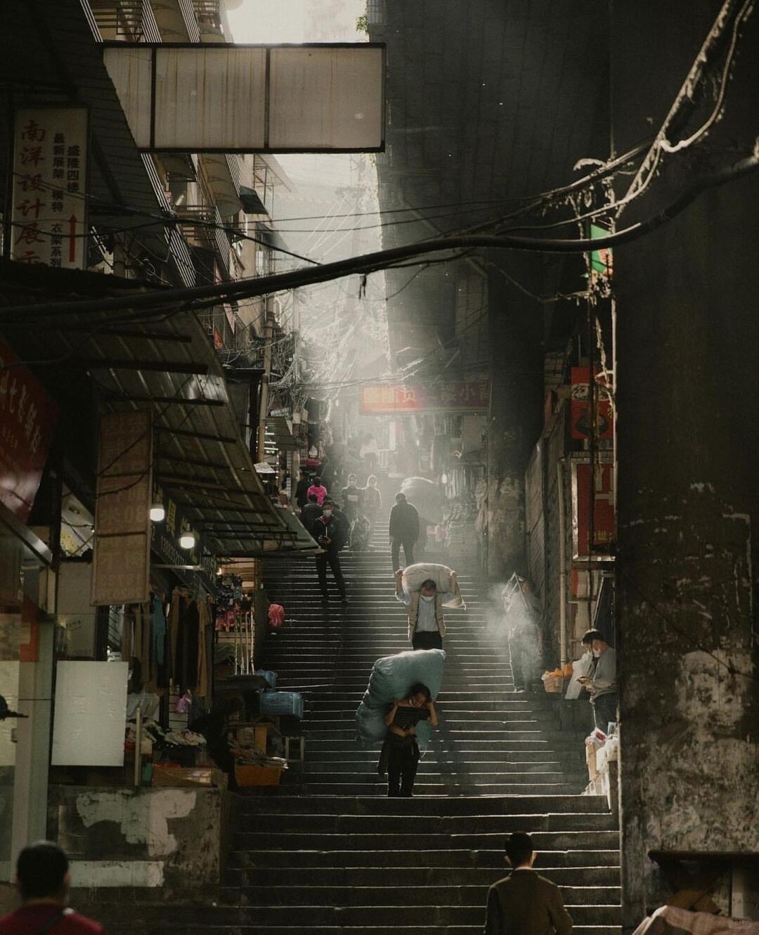 Porters carrying sacks up a steep old market stairway between shopfronts in a misty Chongqing lane at dawn.