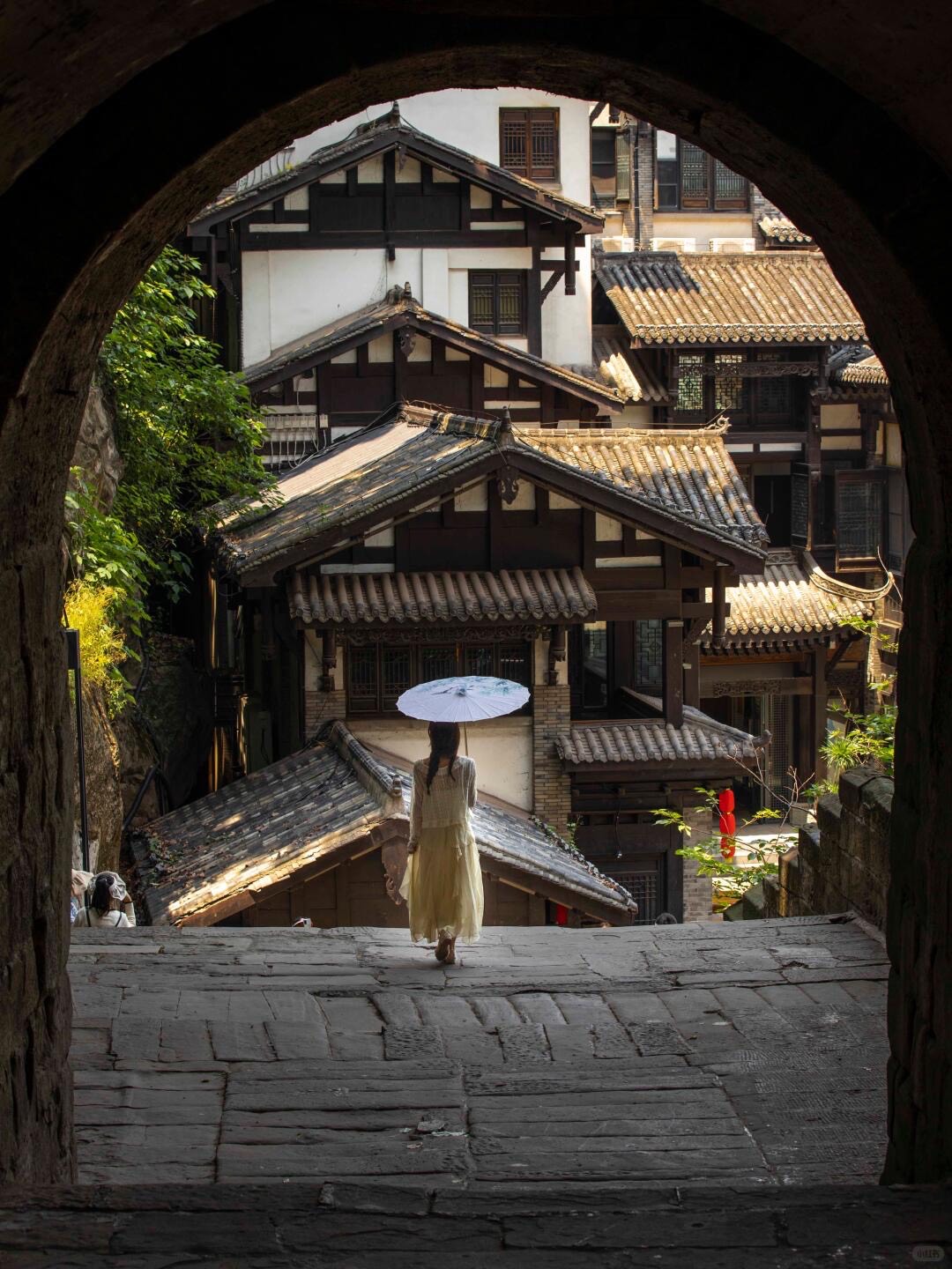 Woman with a paper parasol descends stone steps framed by an arch, facing tiered wooden houses in old Chongqing.