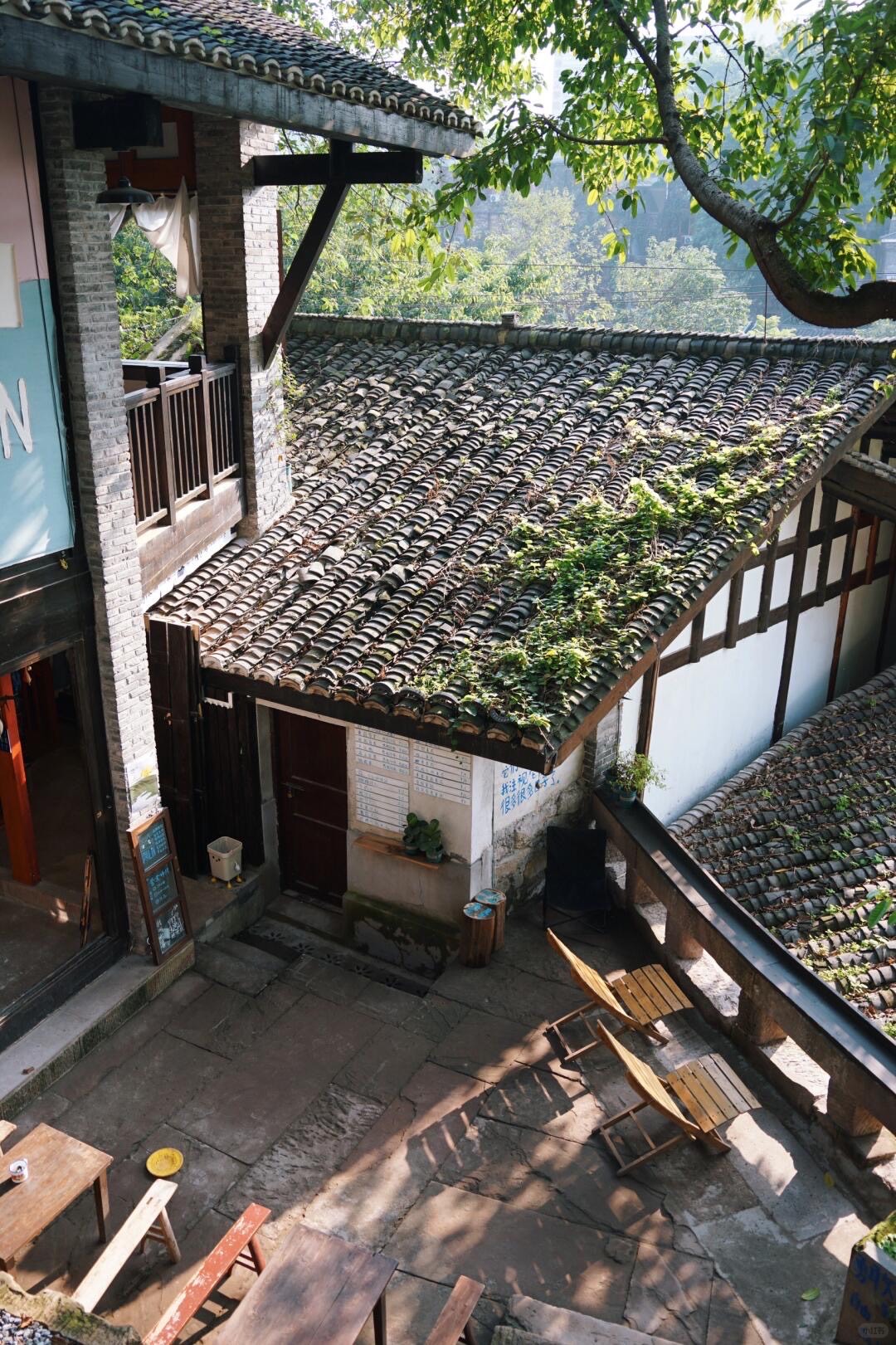 Sunlit stone courtyard with wooden chairs beneath vine-draped grey-tiled roofs of a traditional Chongqing old-town building.