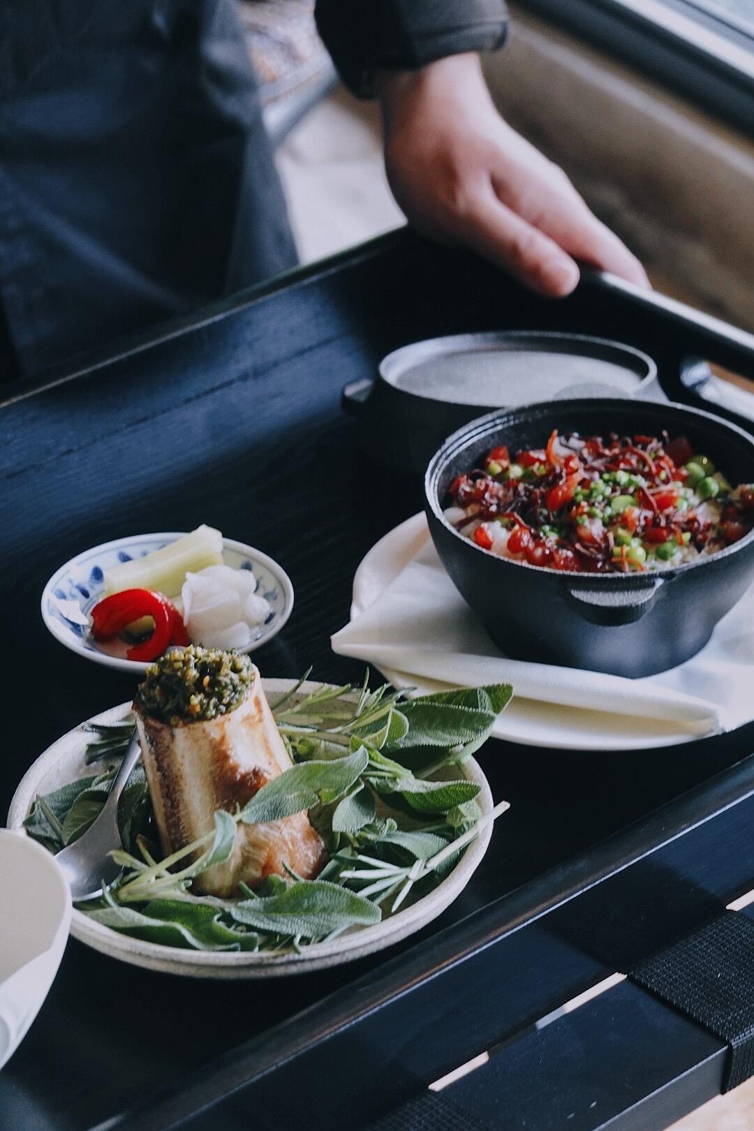 Waiter serving a black tray with a herb-garnished dish, pickles and a claypot of rice at a Chongqing restaurant.