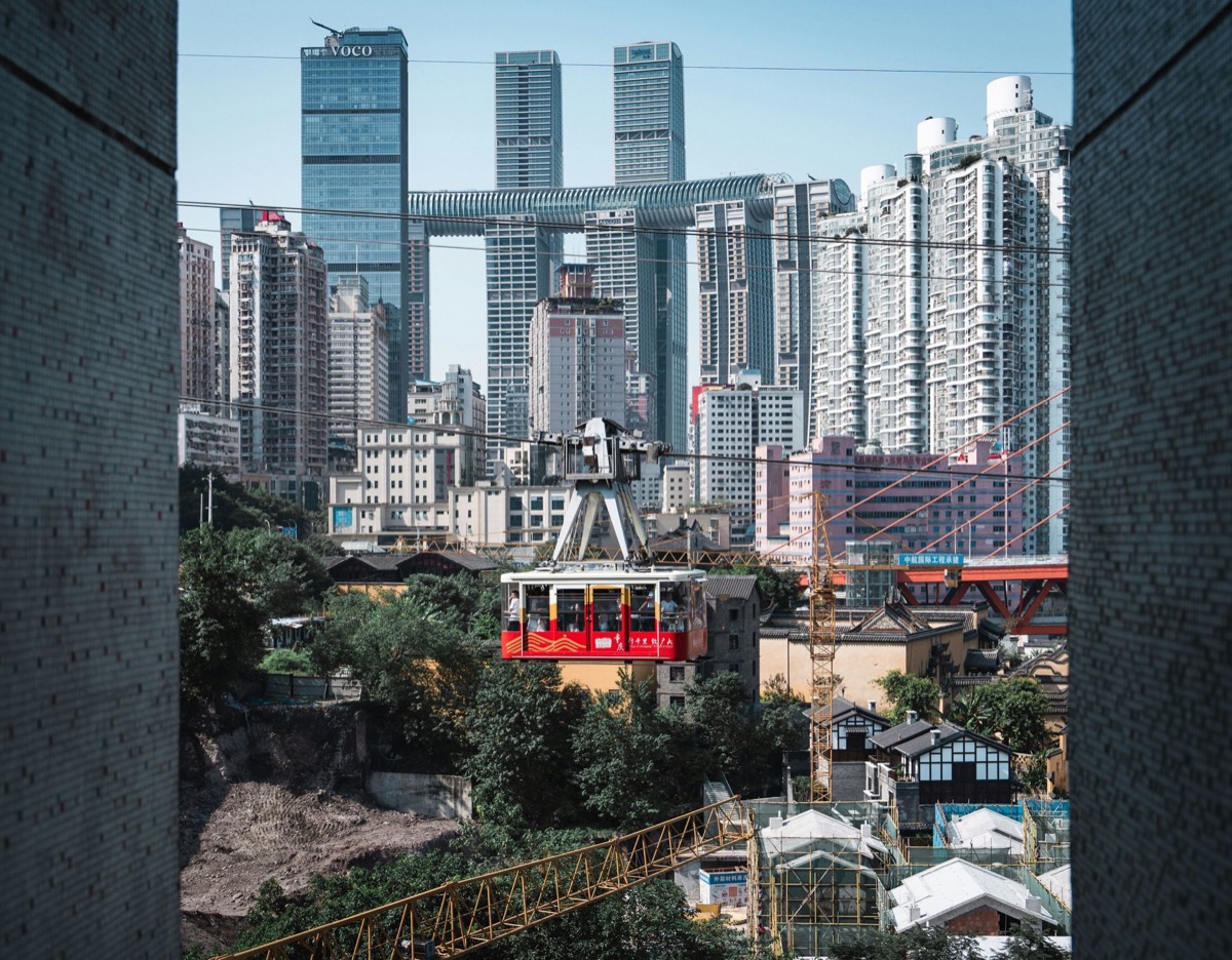 Red cable car crossing above old rooftops with Chongqing's cluster of glass skyscrapers rising behind.