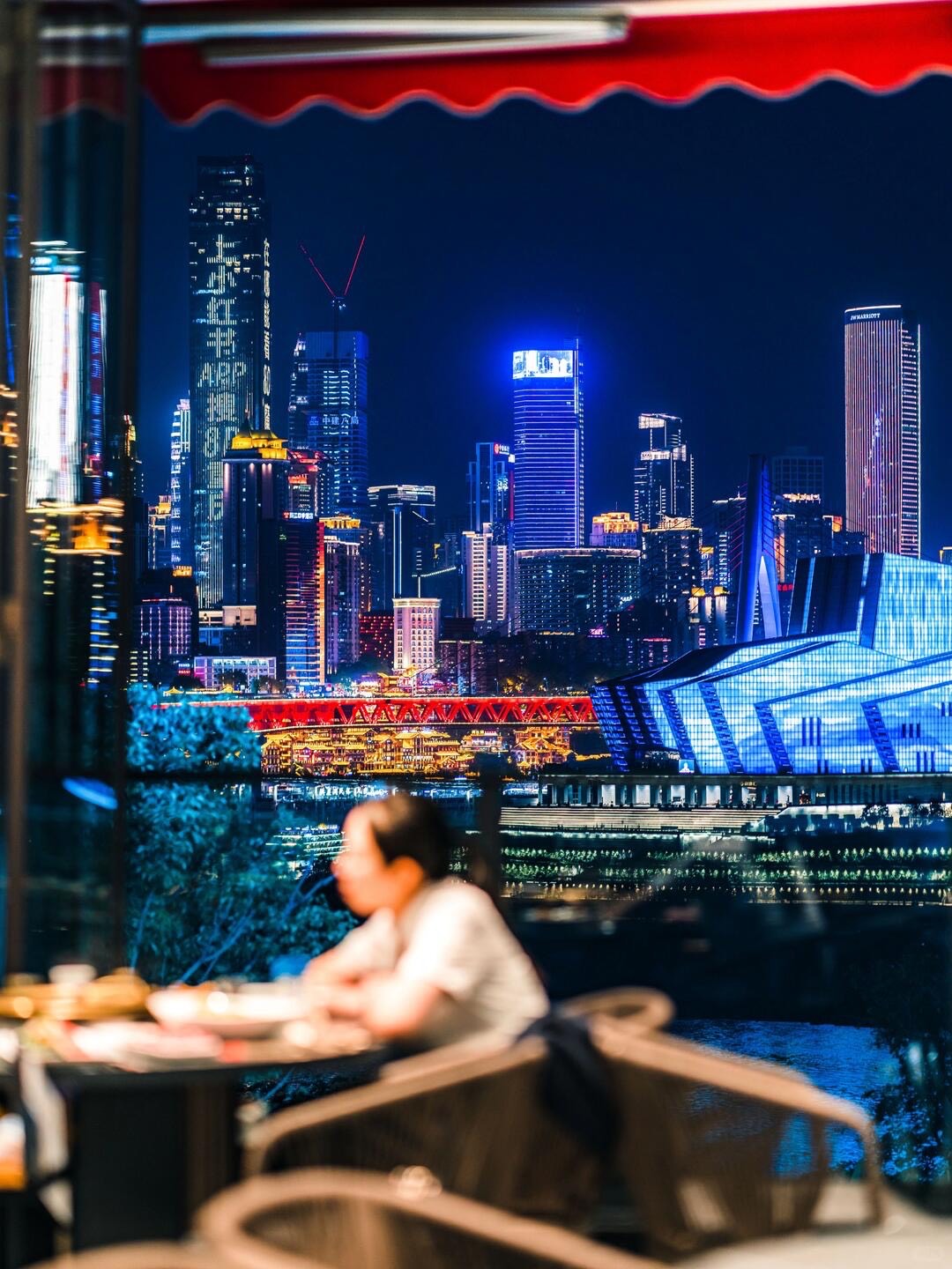 Diner at a riverside terrace restaurant overlooking Chongqing's illuminated high-rise skyline and bridge at night.