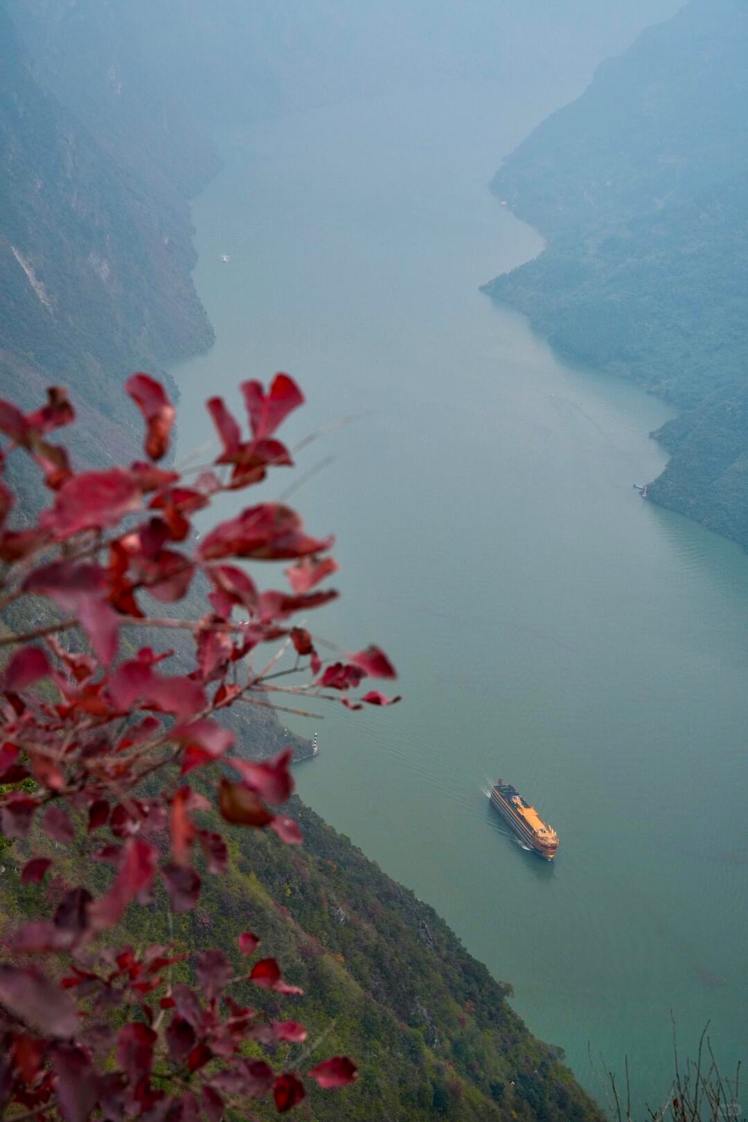 A cruise ship sails a misty jade river through steep Sanxia gorge cliffs, framed by red autumn foliage in the foreground.