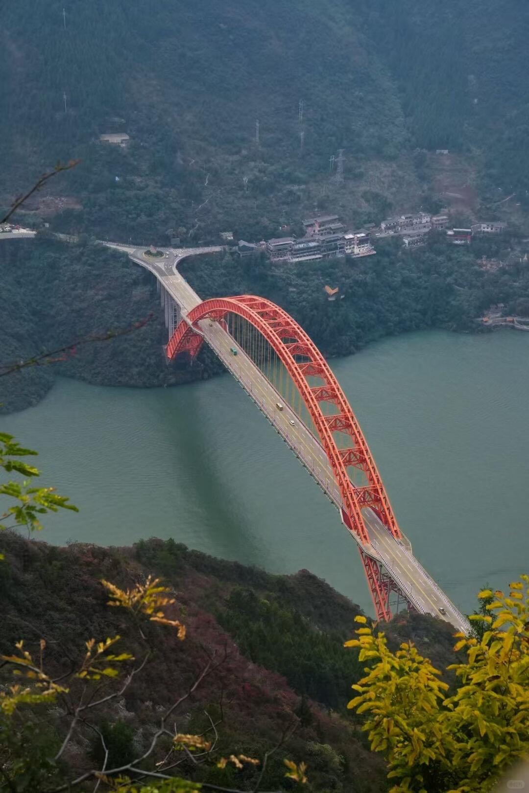 Aerial view of a red steel arch bridge spanning a green river between forested gorge cliffs near Chongqing.