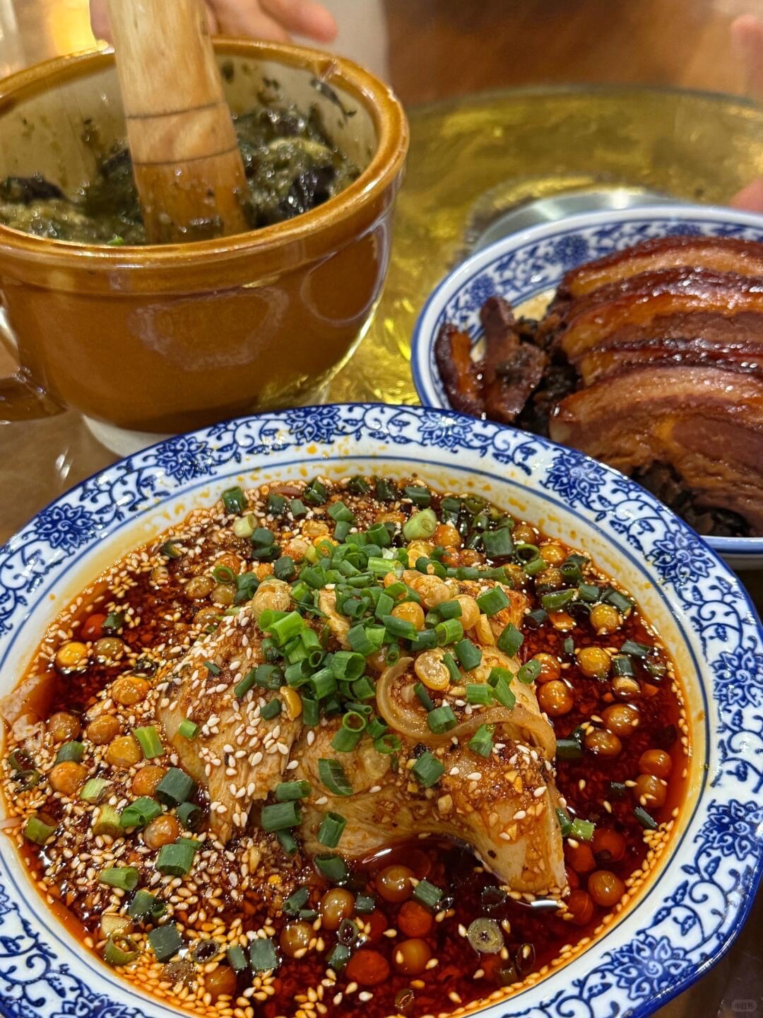 Blue-and-white bowl of sliced chicken in chilli oil with sesame and spring onion beside braised pork and a clay pot.