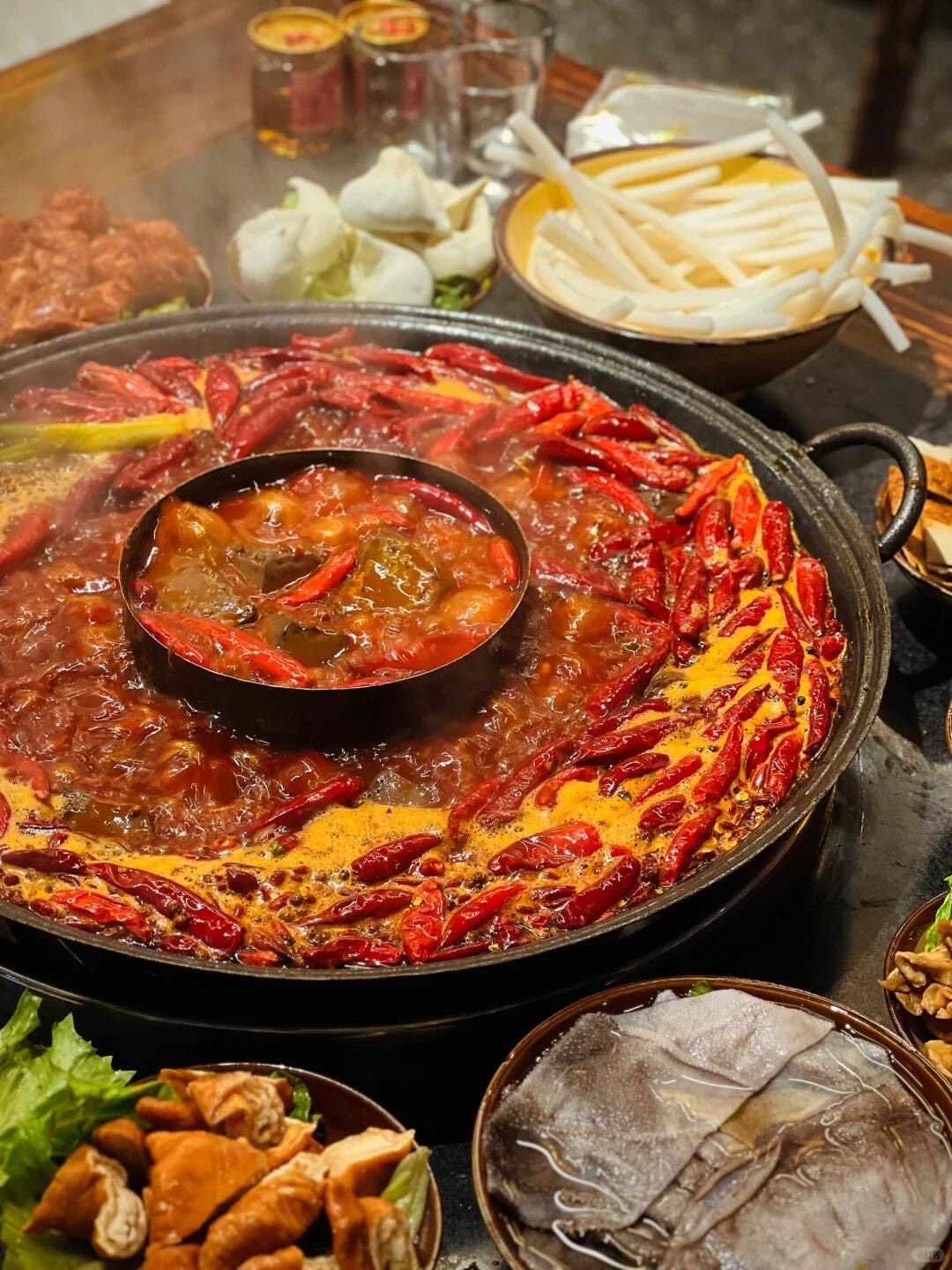 Bubbling chili-laden Chongqing hotpot ringed with dried peppers, surrounded by dishes of ingredients on a table.