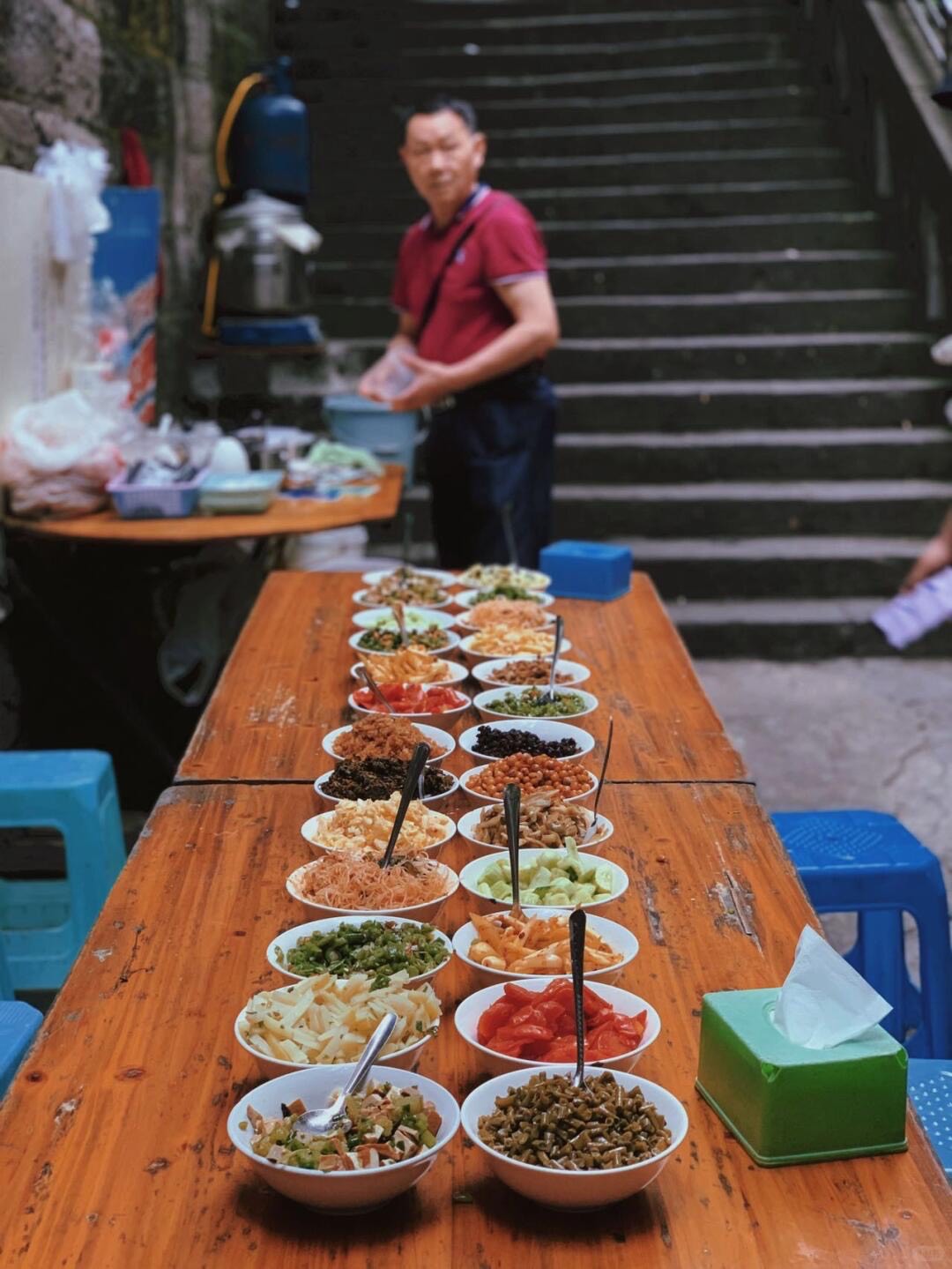 Vendor beside a long table lined with bowls of colourful noodle toppings and pickles at a Chongqing stairway food stall.