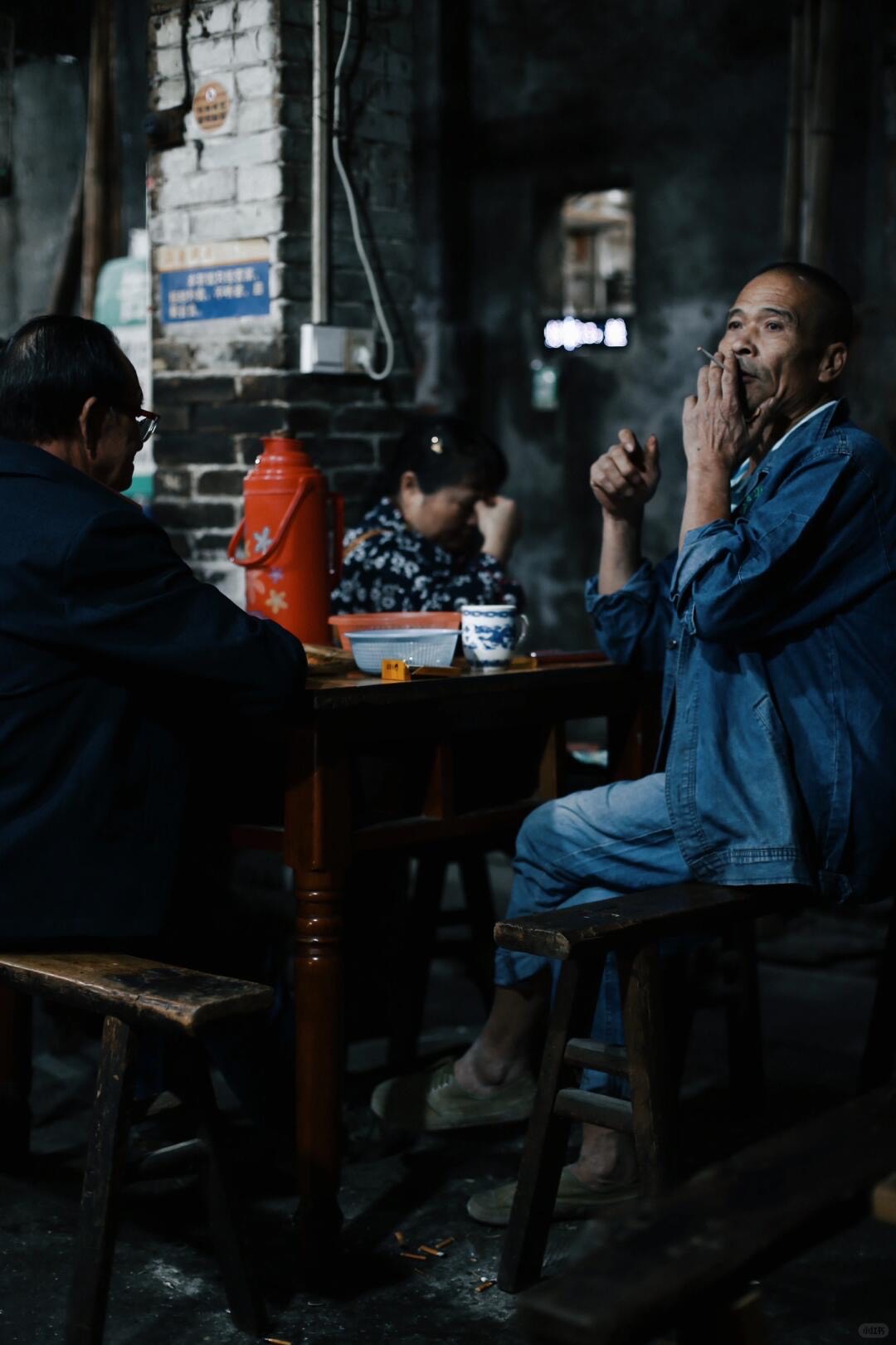 Locals sit on wooden benches around a table in a dim brick-walled teahouse, one man smoking beside a red thermos.