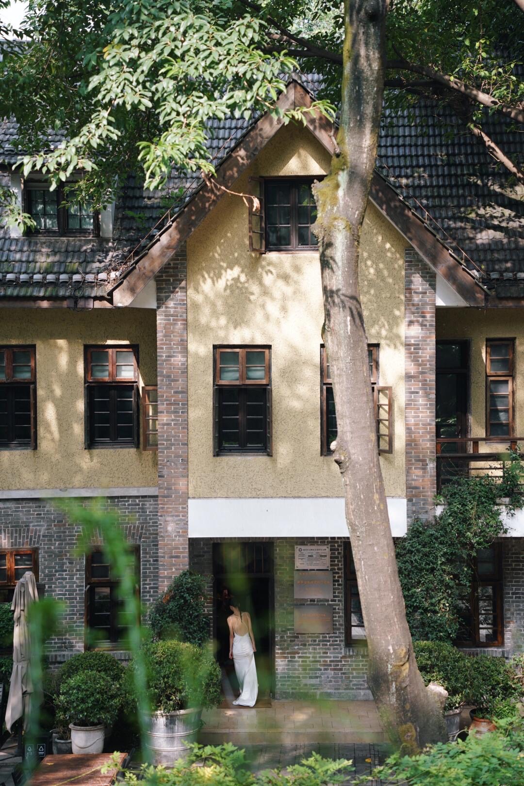 Woman in a long white dress at the doorway of a vintage brick-and-stucco villa framed by a tall tree and garden greenery.