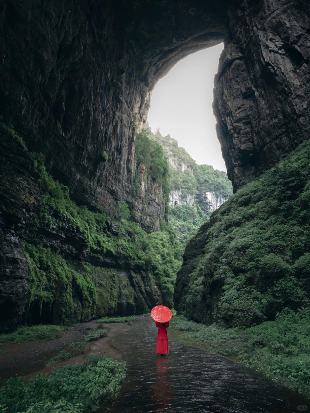 Figure in red with a paper umbrella standing under a colossal natural rock arch in Chongqing's Wulong karst.