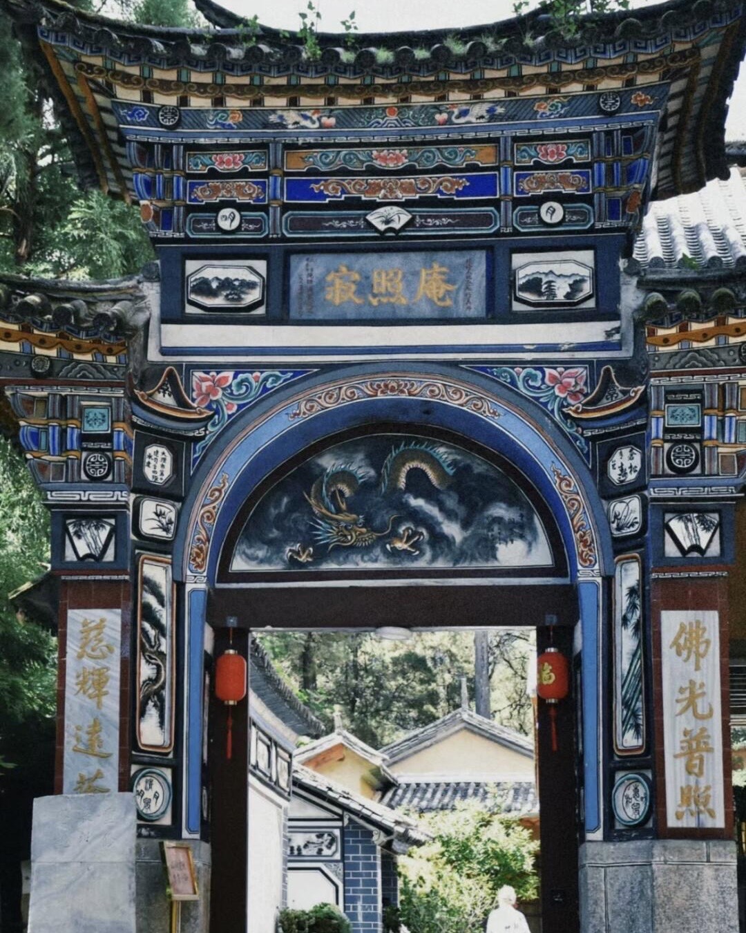 Ornately painted Bai-style temple gateway with a dragon mural and calligraphy panels set against the wooded Cang Mountain foothills near Dali.