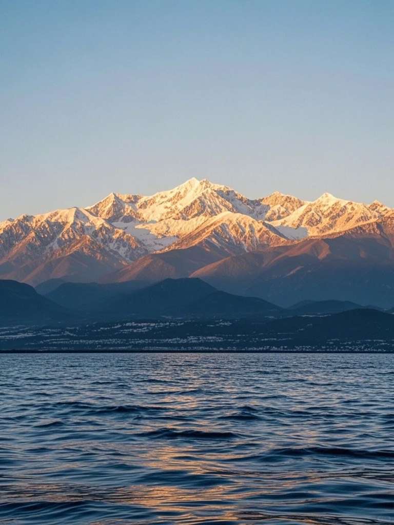 Snow-capped mountains glowing in golden light above the calm waters of Erhai Lake near Dali at sunset.