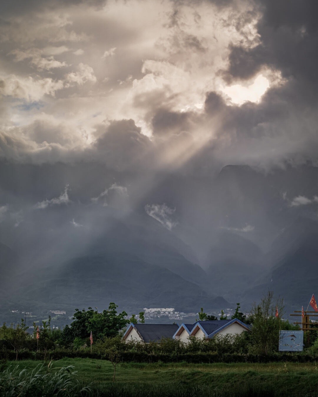 Sunbeams breaking through storm clouds over misty mountains above village rooftops and green fields near Dali's Erhai Lake.