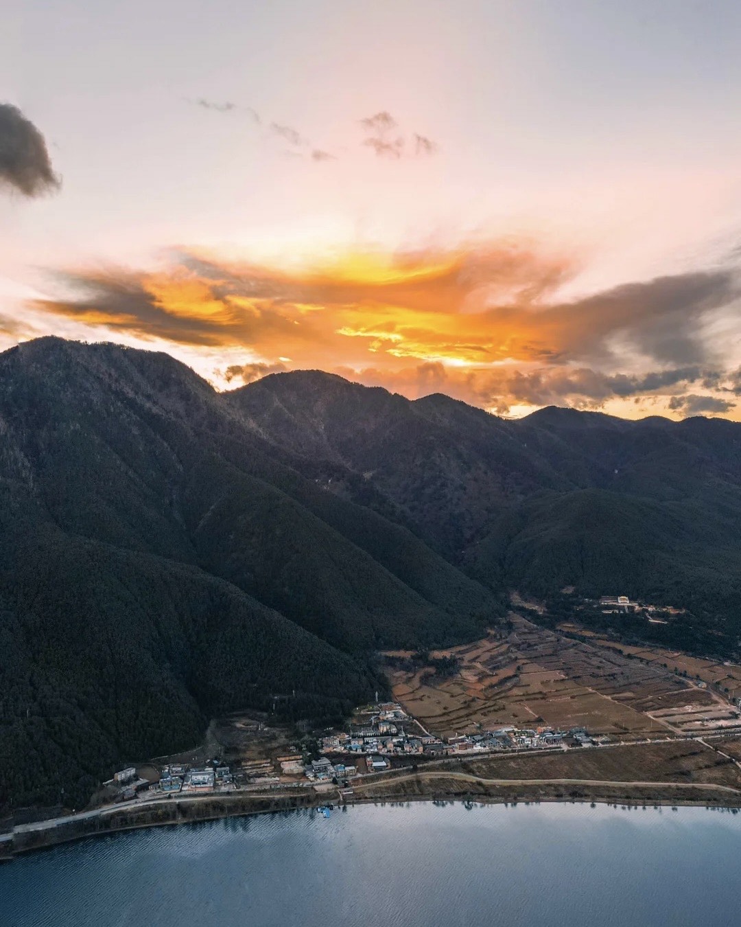 Aerial view of a lakeside village and farmland below forested mountains beside Erhai Lake, under a fiery sunset sky near Dali.