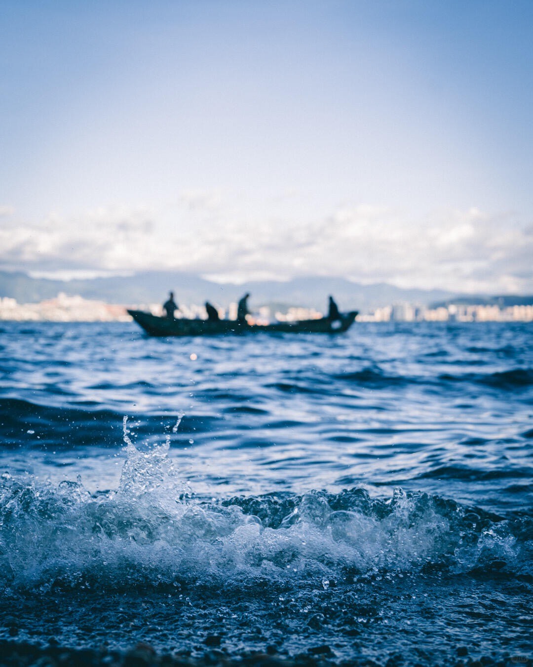 Figures on a wooden boat drifting on Erhai Lake behind splashing waves, with a hazy shoreline and mountains beyond.