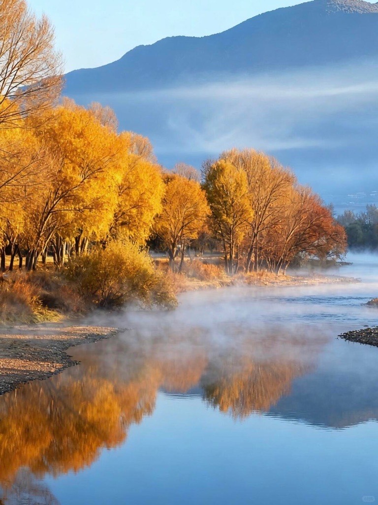 Golden autumn trees reflected in a calm river with rising mist and hazy mountains beyond near Dali.