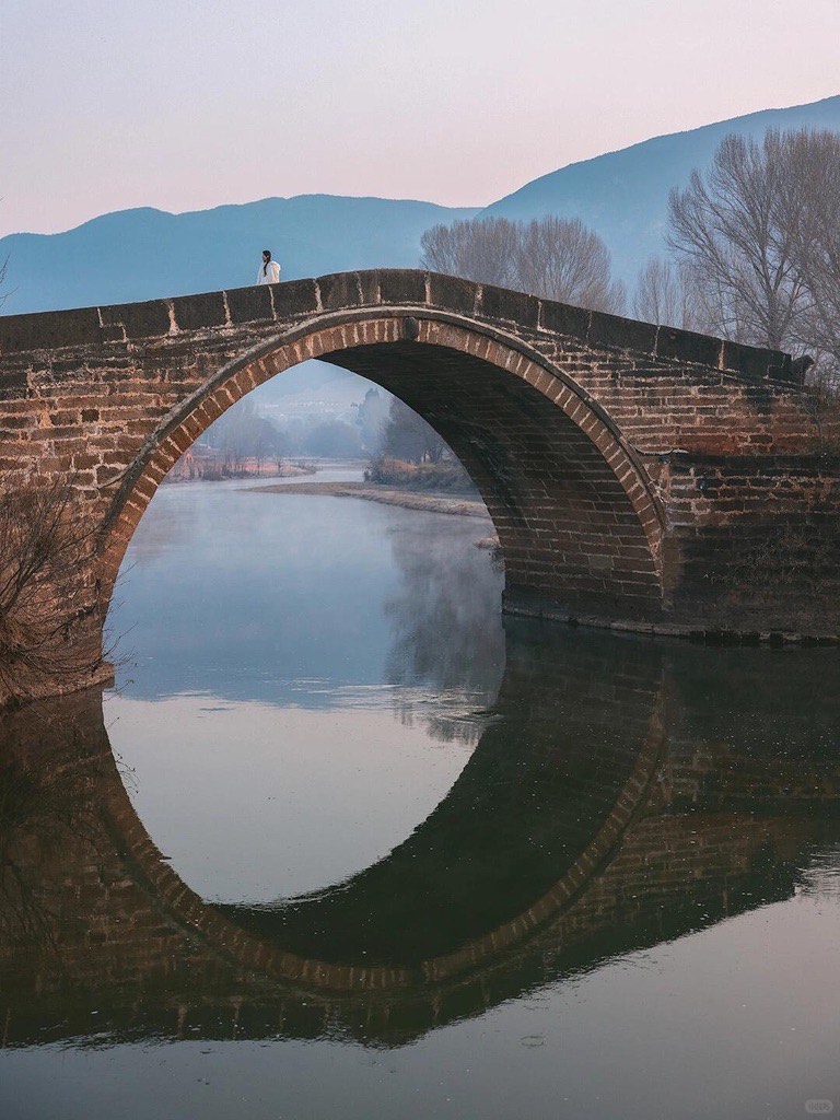 Old stone arch bridge over a still river at Shaxi Ancient Town near Dali, its circle mirrored in the misty water at dusk.