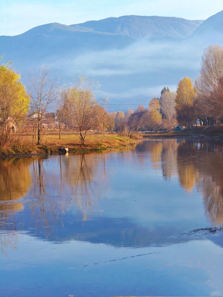 Calm misty river near Shaxi reflecting bare autumn trees with layered mountains under soft morning light.