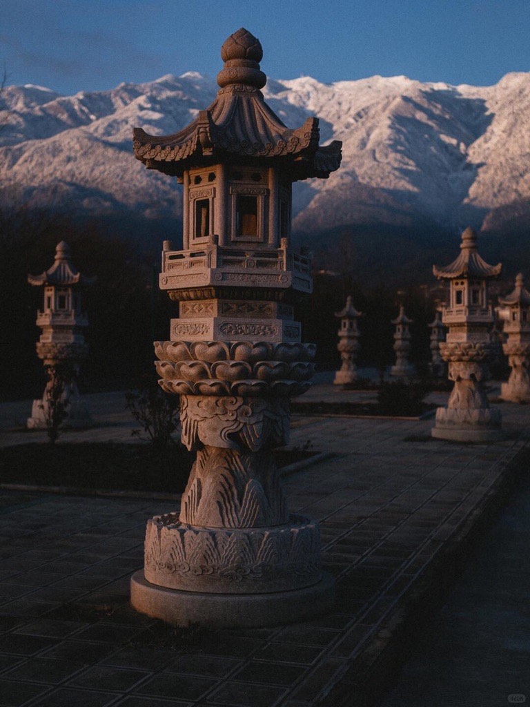 Carved stone pagoda lanterns lining a courtyard at dusk with the snow-dusted Cang Mountain range behind them near Dali.