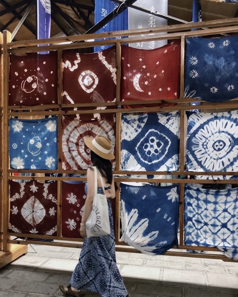 Woman in a straw hat walking past a wooden rack of red and indigo tie-dyed batik fabrics in Dali.
