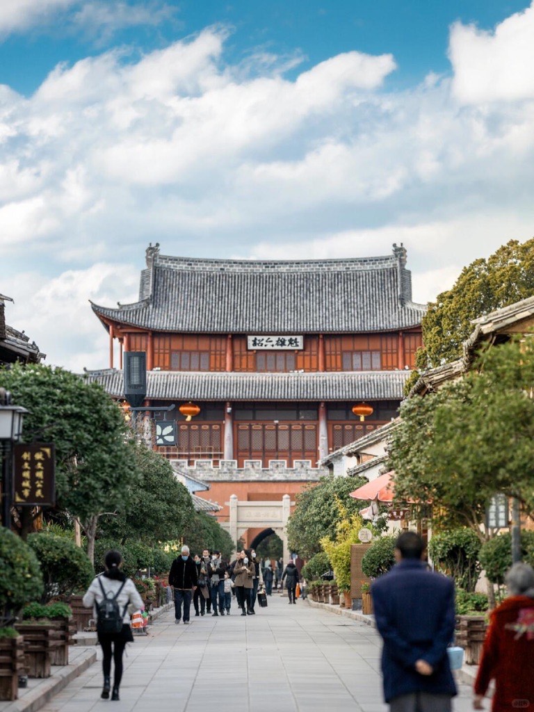 Visitors walking a tree-lined street toward a traditional tiered gate tower in Weishan Ancient Town near Dali.