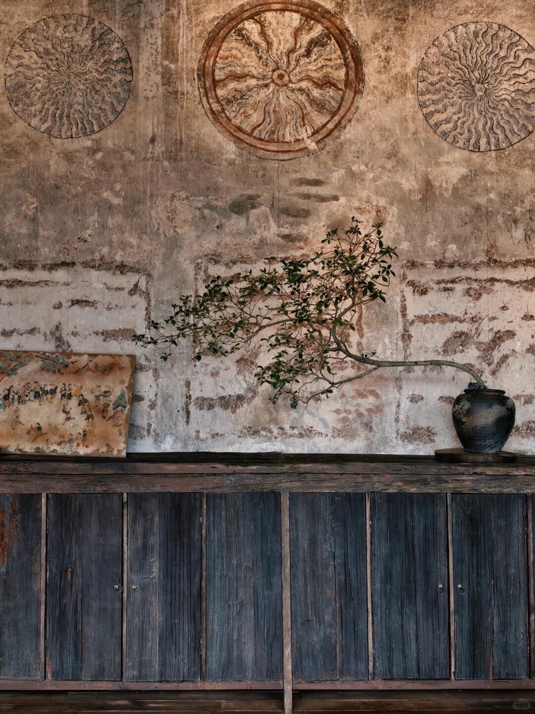 Weathered old wall in Weishan with faded sun-disc murals above a dark wooden cabinet and branch in a black vase.